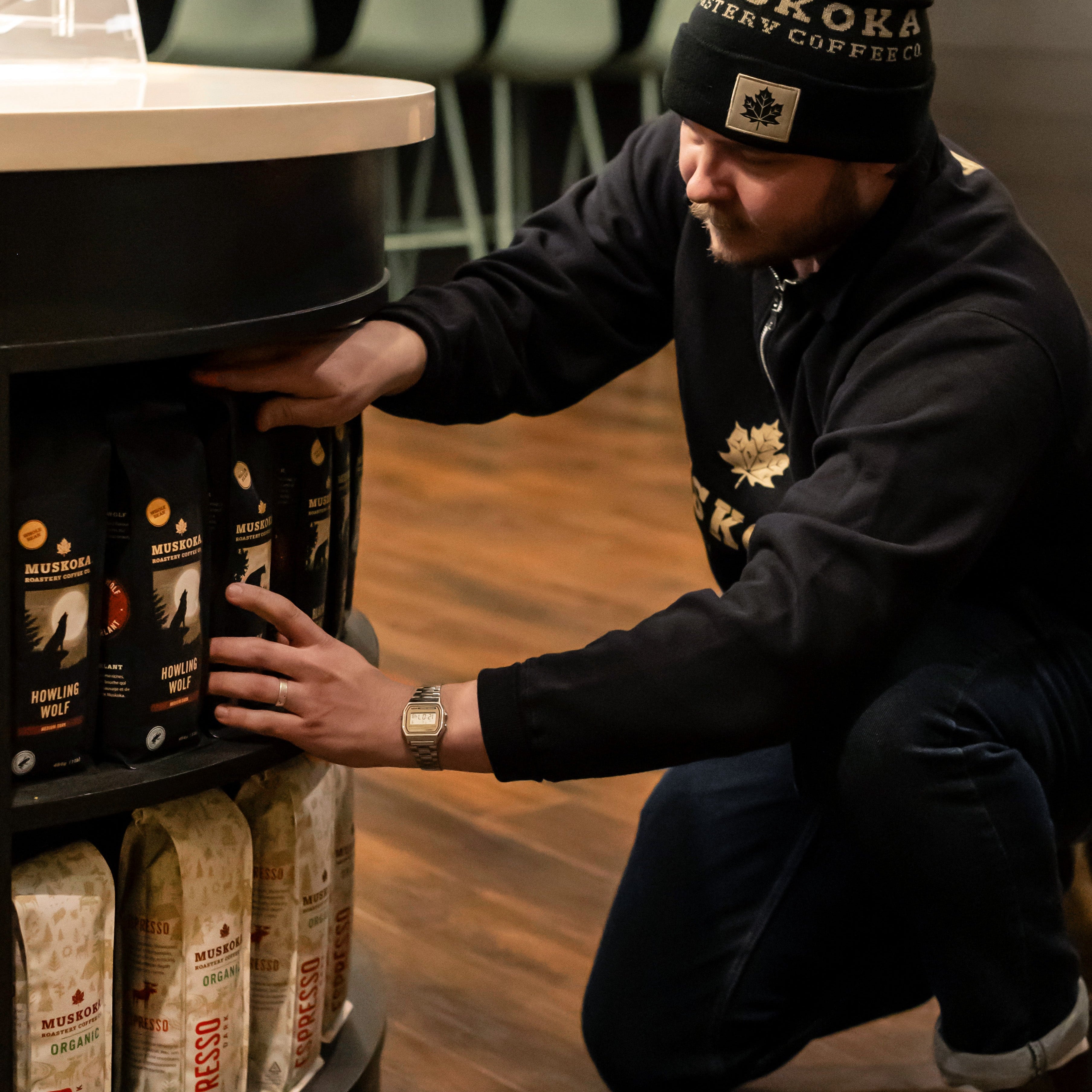 A man in a black hoodie and beanie kneels beside a display shelf, reaching toward several bags of Muskoka Roastery Howling Wolf coffee. The setting appears to be a warm, indoor retail or cafe environment with wood flooring.